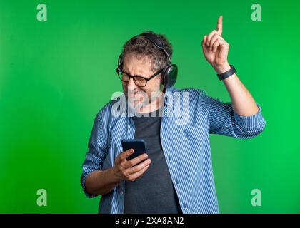 Mature man wearing headphones and a striped shirt, dancing and smiling while holding a smartphone against a green background. The image conveys joy, m Stock Photo