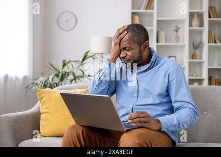 Frustrated man sitting on the couch with a laptop, holding his head. He appears to be dealing with stress or a problem at home. Stock Photo