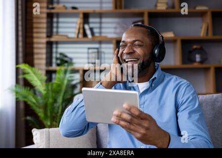 Smiling man sitting at home wearing headphones and using a tablet, enjoying music and relaxation. Stock Photo