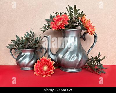Vintage Pewter Teapots with Bright Orange Gerbera Flowers and Greenery on Red Surface Stock Photo