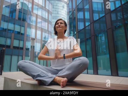 De-stress, unwind idea. peaceful female freelance employee practicing yoga exercises while sitting in bunch under glass skyscrapers after hard working Stock Photo