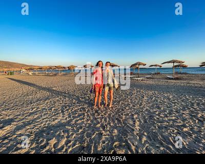 Two women in swimsuits strolling on the beach with umbrellas in Greece Stock Photo