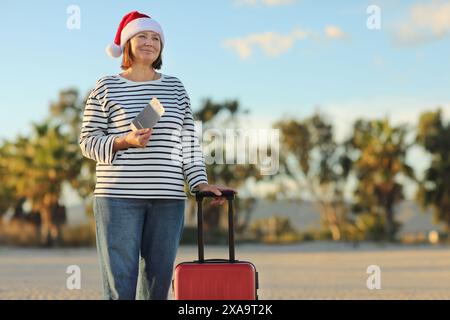 Independent mature woman in Christmas Santa Claus hat, striped shirt holds passport, boarding ticket, suitcase on seashore with palms backdrop celebra Stock Photo