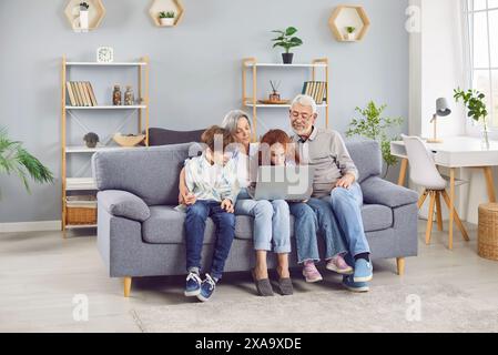 Smiling seniors resting on couch with grandchildren at home enjoying weekend using laptop. Stock Photo