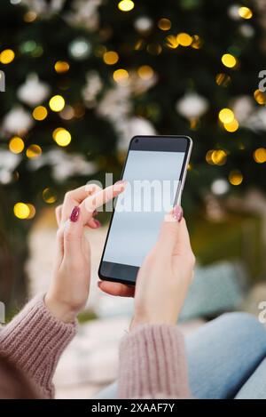 christmas mockup hands holding phone with empty screen on background of beautiful christmas tree with lights. Stock Photo