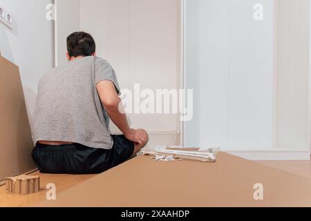 Young man on his back sitting on the floor assembling a white bedroom cabinet Stock Photo