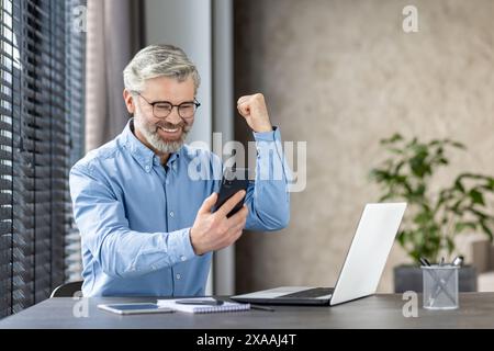 Happy senior businessman celebrating success while video calling on a smartphone at his home office with a laptop and documents. Stock Photo