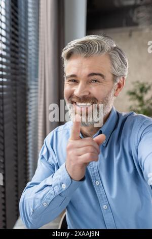 Middle-aged man in blue shirt smiling and pointing while video calling from home office, expressing happiness and engagement. Stock Photo