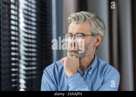 Mature man with glasses looking thoughtful while standing by a window in a bright room. Reflective and contemplative expression Stock Photo
