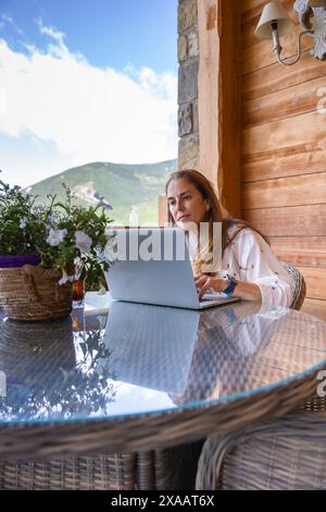 mature Caucasian woman teleworking with laptop, located outside an open-air house with mountains in background Stock Photo