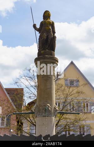 Feuchtwangen, Germany - June 6, 2023: A stone statue of a female figure holding a spear atop a fountain in a German town. The statue is decorated with Stock Photo