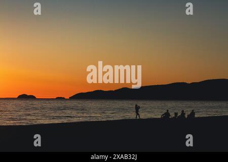 While watching the evening sunset on the shores of Agawa Bay campground a silhouetted young woman takes a photograph of her family while they relax Stock Photo