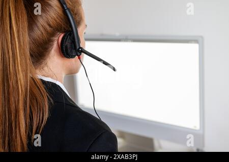 Rear view of call centre female worker in headset in front of computer display with blank screen. Stock Photo