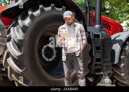 Man farmer stands next to an agriculture tractor and using his digital tablet. Agricultural mechanic searching information on the internet. Stock Photo