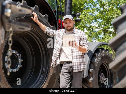 Man farmer stands next to an agriculture tractor and using his digital tablet. Agricultural mechanic searching information on the internet. Stock Photo