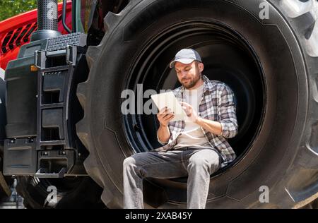 Man farmer sits on the big wheel of agricultural tractor and using his digital tablet. Stock Photo