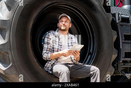 Man farmer sits on the big wheel of agricultural tractor and using his digital tablet. Stock Photo