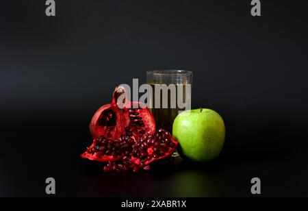 A tall glass of freshly squeezed fruit juice against a black background, next to a broken pomegranate fruit with seeds and a ripe green apple. Close-u Stock Photo