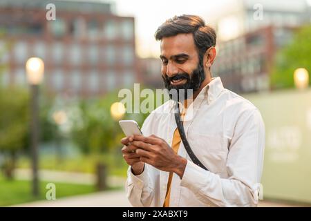 Man Smiling While Using Smartphone in Urban Park at Sunset Stock Photo