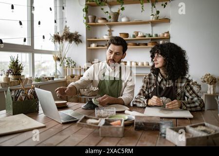 Bearded man in apron showing pottery techniques to woman while sculpting clay in creative workshop using online video tutorial. Stock Photo