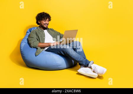 Portrait photo of young funny mexican guy student remote studying in khaki shirt sitting pouf at lounge isolated on yellow color background Stock Photo