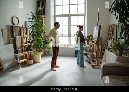 Artist spouses engaged in creative process together, husband accompanies wife working on painting Stock Photo