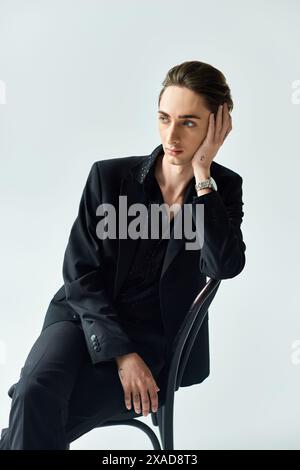 A stylish young queer person in a black suit confidently sits on a chair in a studio, exuding pride and elegance. Stock Photo