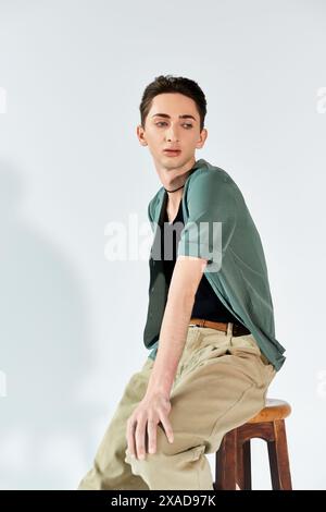 A young man confidently sits on a stool in a stylish green shirt and tan pants against a grey studio backdrop. Stock Photo