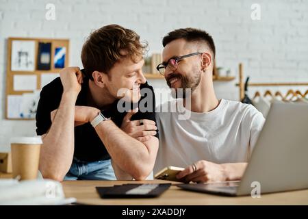 Two men, a gay couple, sit at a table together, focused on a laptop screen in a designer workshop. Stock Photo