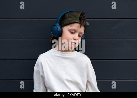 Portrait of a pre-teen boy in a cap with headphones wearing a white t-shirt against a background of wooden planks in graphite color Stock Photo