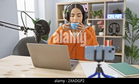Hispanic woman sips coffee while recording in a modern radio studio, symbolizing a casual podcast atmosphere. Stock Photo