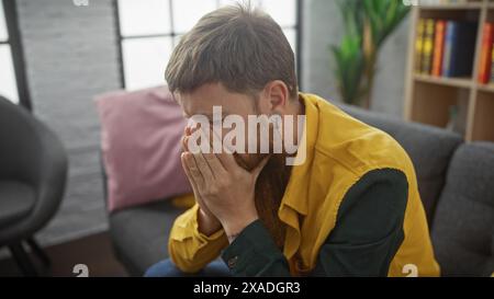 A distraught bearded redhead man sitting indoors, covering face with hands in apparent distress or sadness. Stock Photo