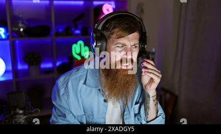 Staunch young redhead man streamer, seriously immersed in digital gaming world, donning headphones in the dimly lit gaming room Stock Photo
