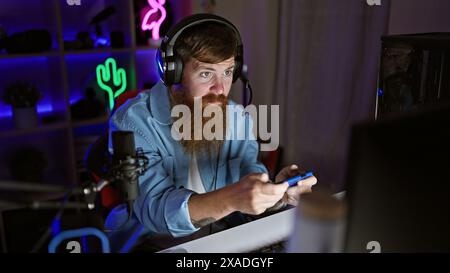 Intensely concentrated young redhead man steaming video game on smartphone in dark gaming room at night Stock Photo