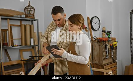 A man and woman in a workshop evaluating a wooden plank while using a tablet. Stock Photo