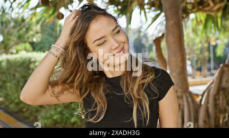 Radiantly confident young hispanic woman joyfully combing hair by hand in a green park, casting a cheerful smile to the side while relishing the fun o Stock Photo