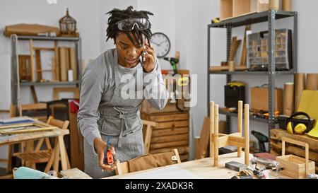 African american woman carpenter using tape measure in a woodworking workshop while talking on the phone. Stock Photo