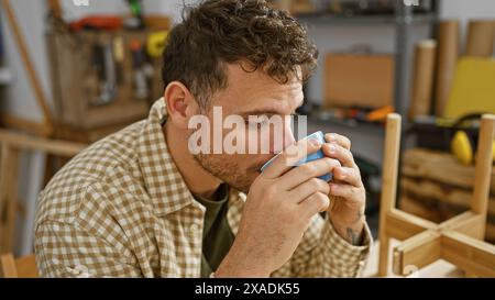 A young hispanic man with a beard enjoys a coffee break in a woodwork studio, reflecting craftsmanship and a relaxed atmosphere. Stock Photo