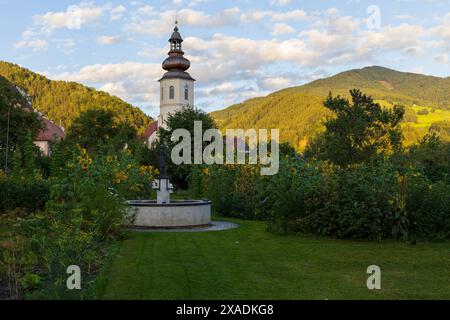 Photo of a medieval bell tower and red roof houses surrounded by green garden with Alpine mountains on a background, alpine village landscape in a sun Stock Photo
