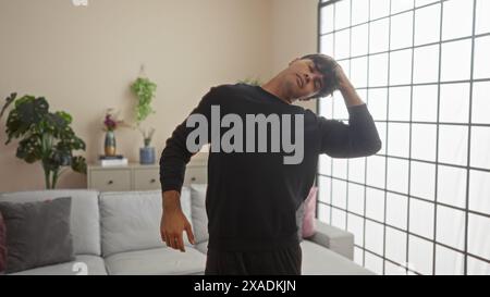 Handsome young hispanic man stretching his neck indoors in a contemporary living room, creating a relaxed and serene atmosphere. Stock Photo