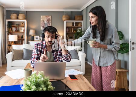 Young woman bringing cup of coffee for her boyfriend while he working online on laptop at table at home. Freelance business man having video call with Stock Photo