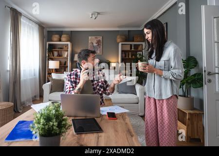 Young woman bringing cup of coffee for her boyfriend while he working online on laptop at table at home. Freelance business man having video call with Stock Photo