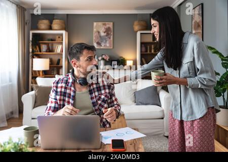 Young woman bringing cup of coffee for her boyfriend while he working online on laptop at table at home. Freelance business man having video call with Stock Photo