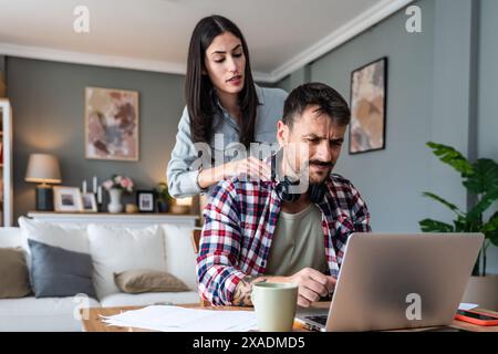 Young stressed freelance man working at home on laptop having neckache and woman give him massage to reduce stress. Male businessman feeling shoulder Stock Photo
