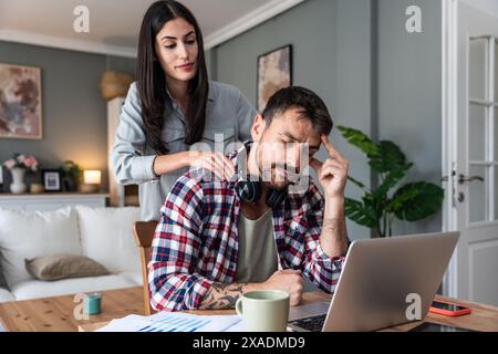 Young stressed freelance man working at home on laptop having neckache and woman give him massage to reduce stress. Male businessman feeling shoulder Stock Photo