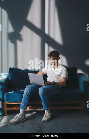 Full length photo of caucasian man working with laptop while sitting on couch in living room. Remote work from home concept. Stock Photo