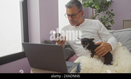 Middle-aged hispanic man holding a small chihuahua dog while examining a medicine bottle near a laptop in a cozy living room. Stock Photo