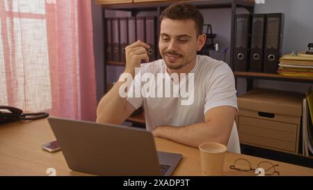 Handsome hispanic man in an office working on a laptop with a coffee cup nearby, capturing a professional indoor setting. Stock Photo