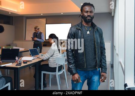 A African Man with dreadlocks and in a black jacket is standing next to the window in the office and behind him are colleagues, some people meeting an Stock Photo