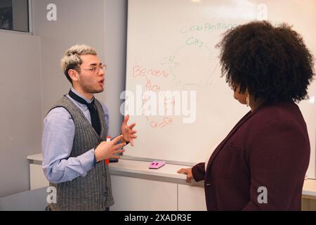 A young Man and a Woman are standing in front of a whiteboard with chemistry formulas written on it. The man is talking to the woman, and they seem to Stock Photo